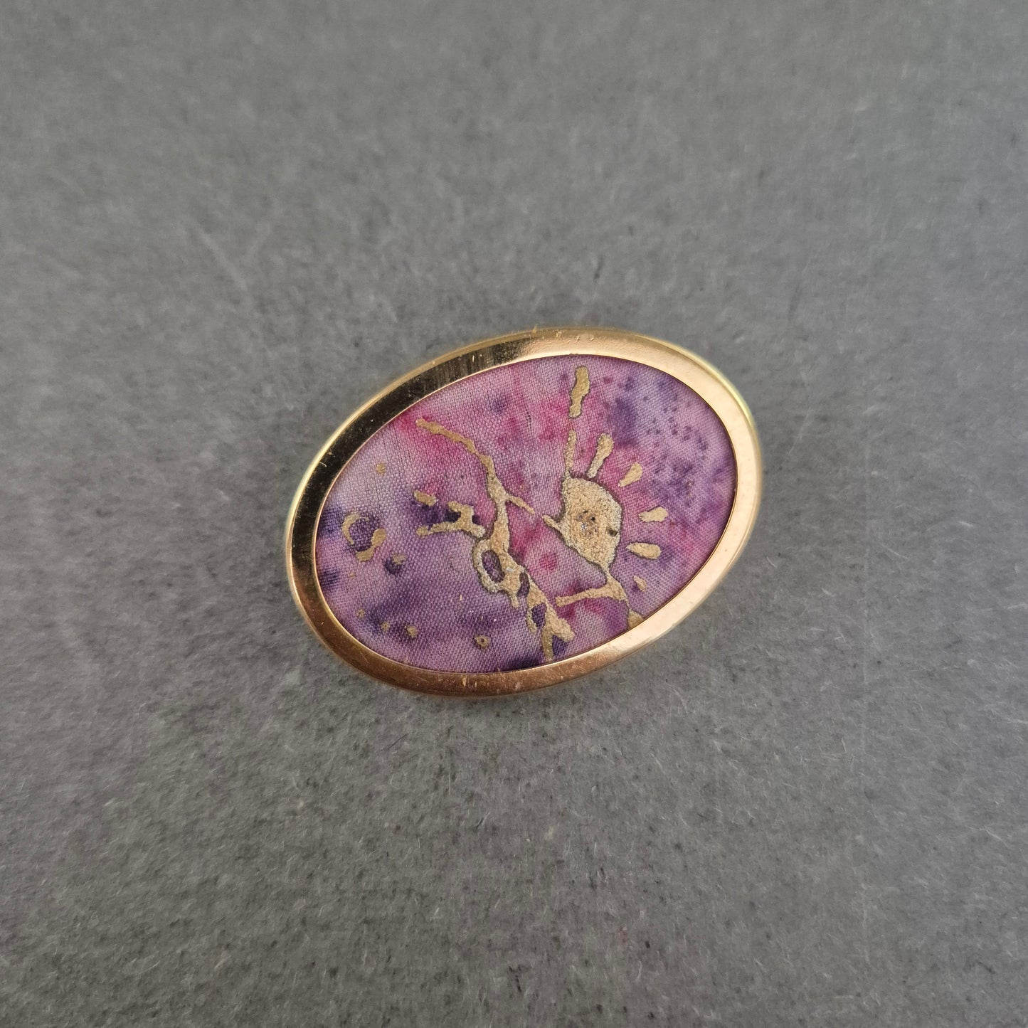 Ovale Brosche mit lila Stoffeinlage und goldener Handzeichnung