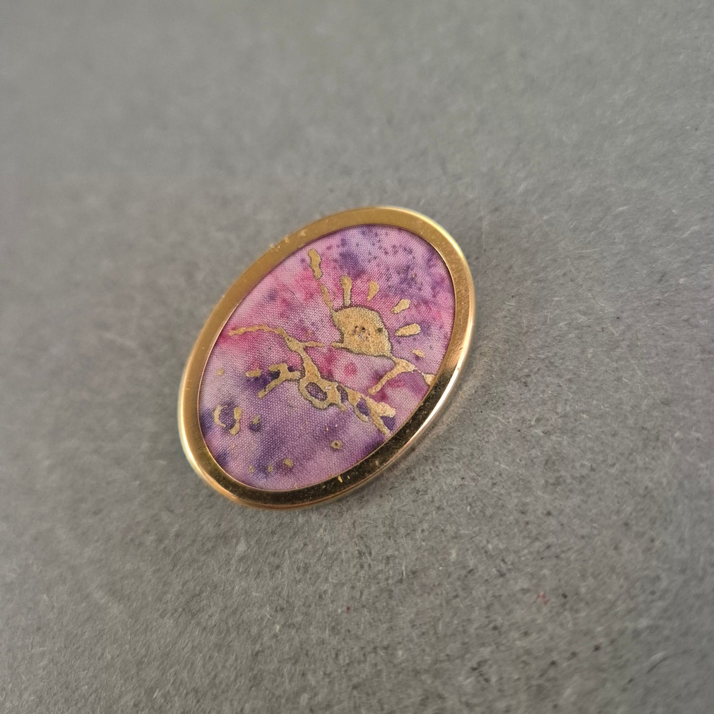 Ovale Brosche mit lila Stoffeinlage und goldener Handzeichnung