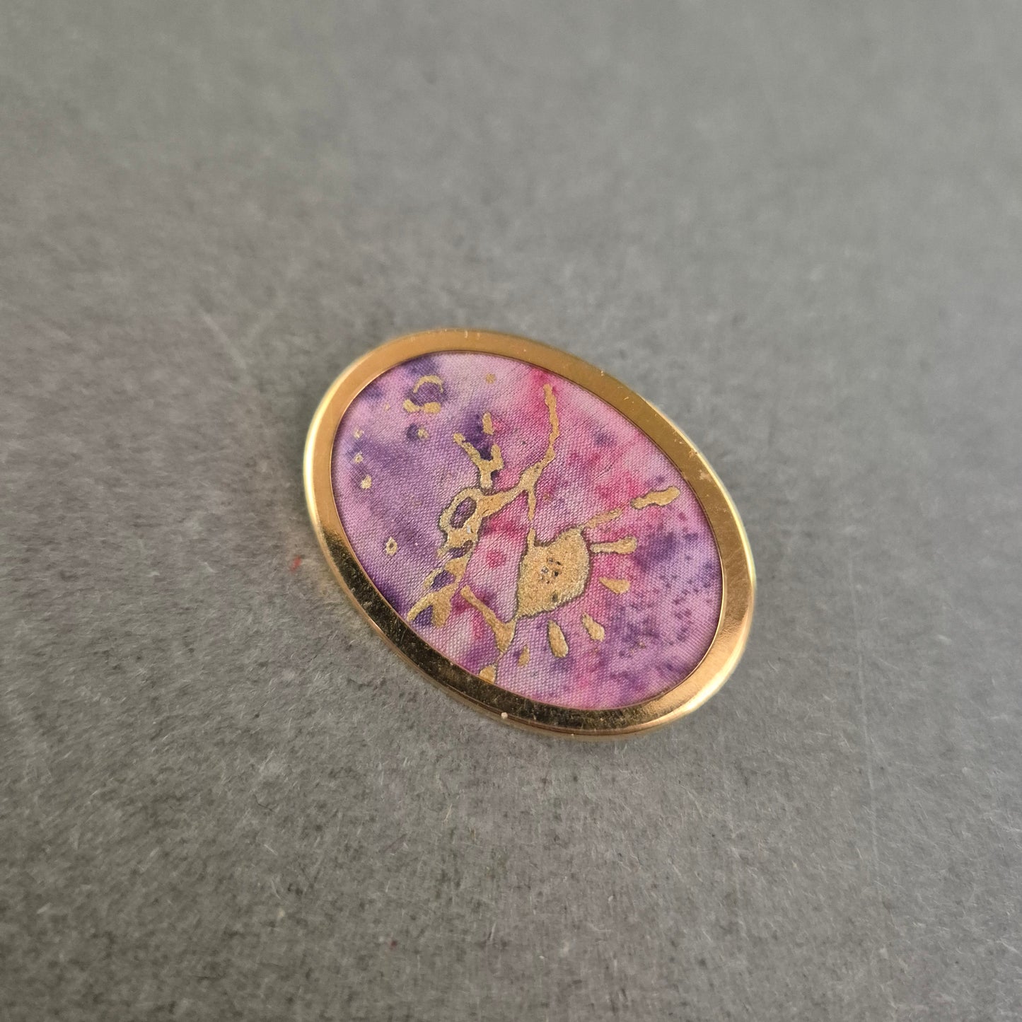 Ovale Brosche mit lila Stoffeinlage und goldener Handzeichnung
